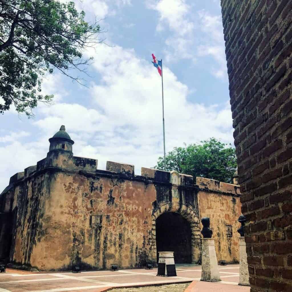 A brown stone fort in the city of Santo Domingo with a tall flag pole in the middle.