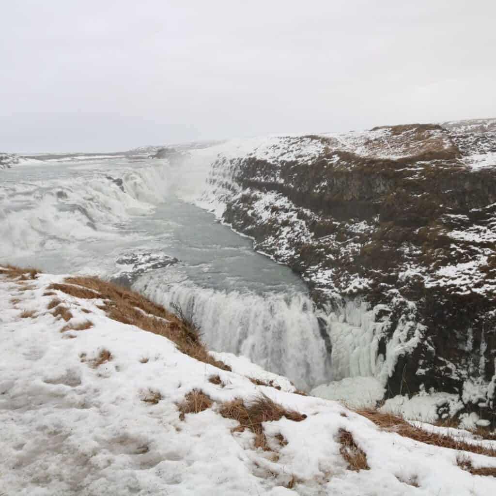 A rushing bluish gray waterfall tumbling over the rocks with snow on the ground in the foreground.