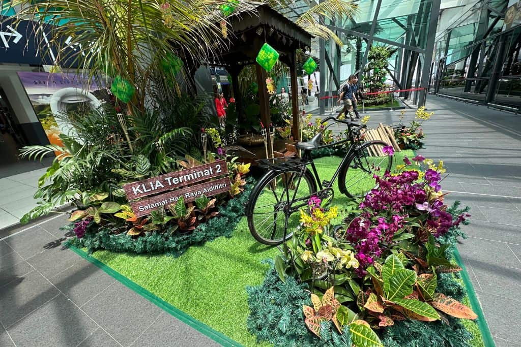 The entrance between the airport terminal buildings that is covered from the hot sun and rain. There is a bicycle with flowers and a sign welcoming you tot he terminal building.