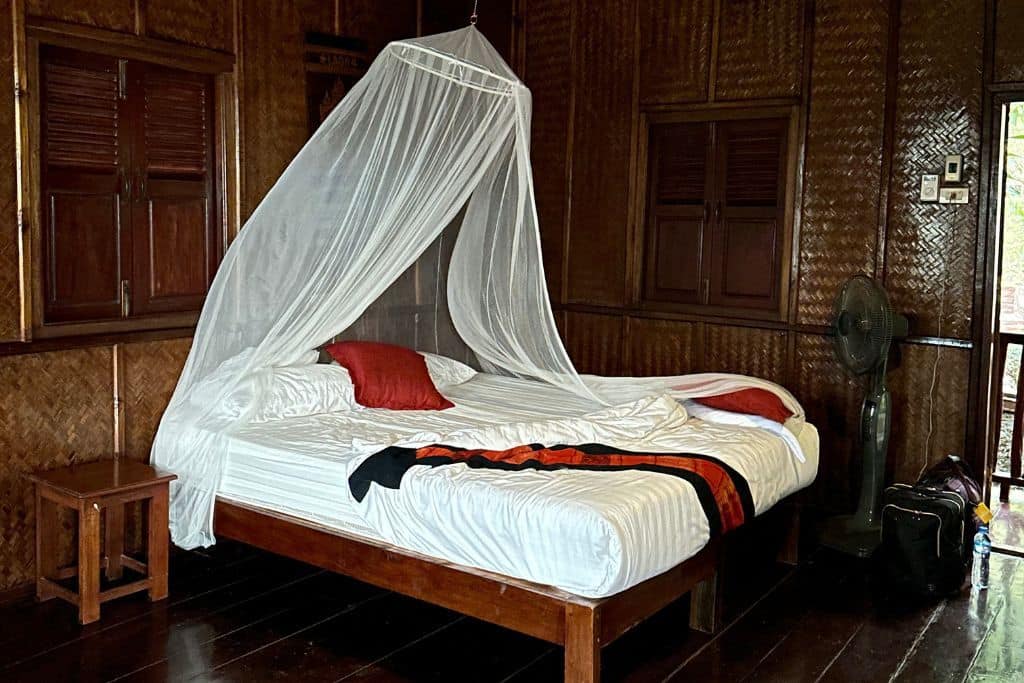 The white covered bed with a mosquito net in a very dark paneled room at the Mekong Riverside Lodge