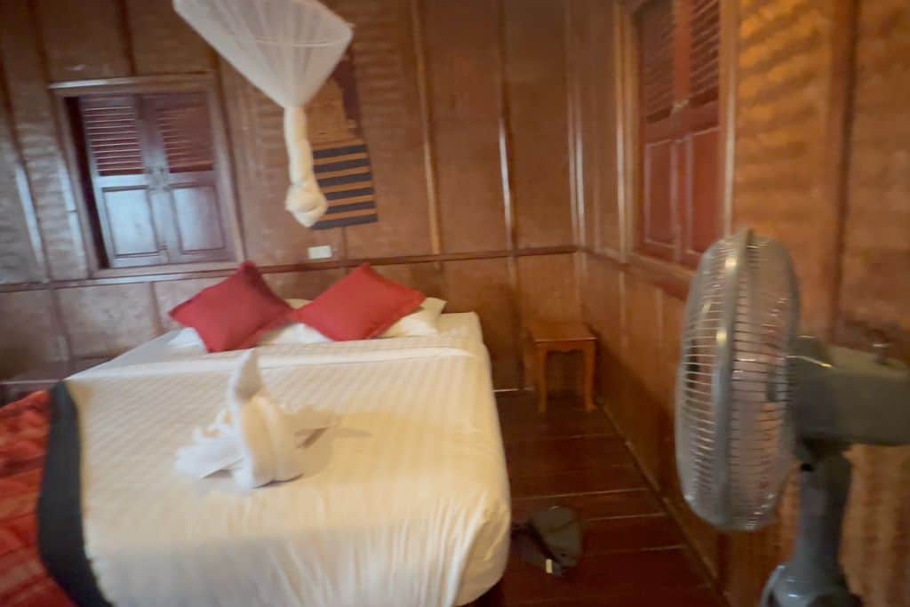 The white covered bed in a dark paneled room with a standing fan near the foot of the bed at the Mekong Riverside Lodge