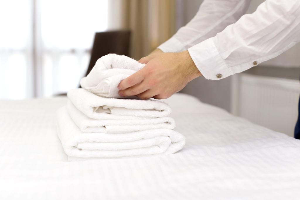 A member of the cleaning staff folding towels on a white bed