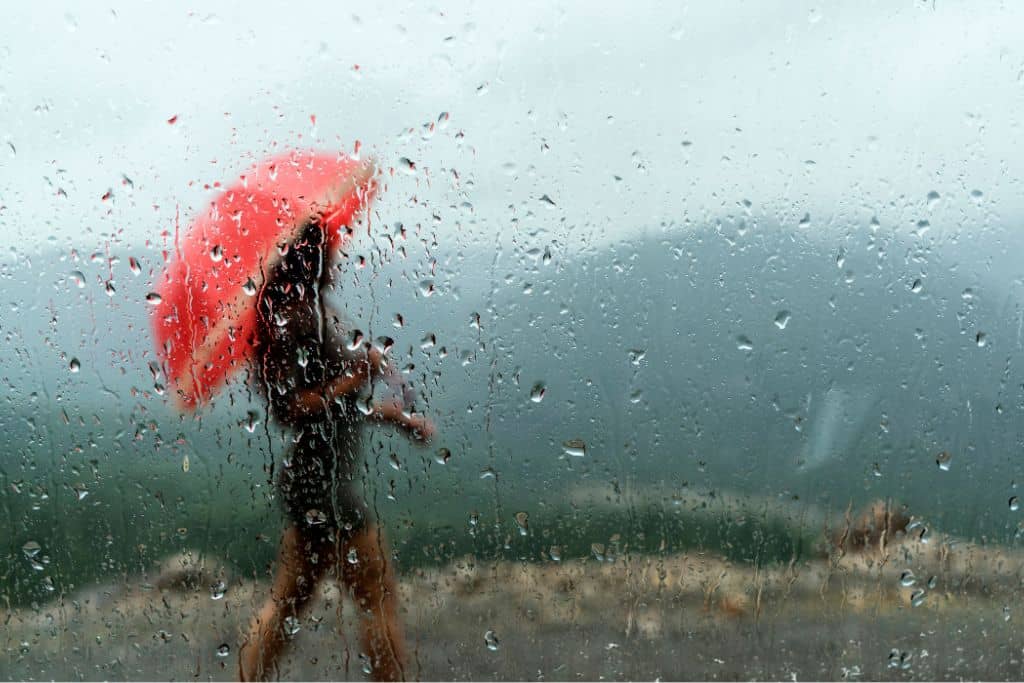 A young woman walking in the rain with a red umbrella with green mountains in the background.