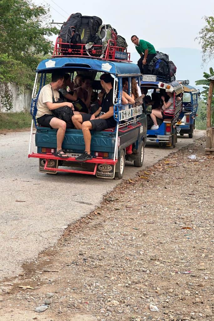 A crowded songtheaw packed full of passengers with luggage stacked on top. The vehicle is open sided with benches along the sides for seating.