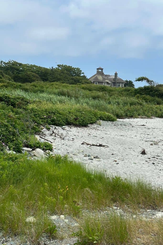 A large mansion seen in the distance with a sandy beach and green shrubs leading down tot he water.