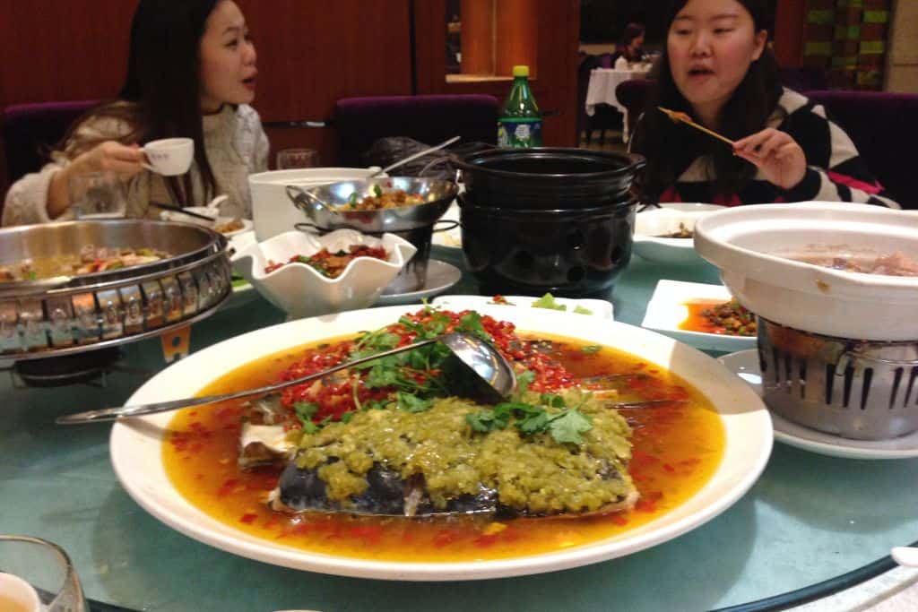 A whole cooked fish with peppers and assorted chopped vegetables on top served on a large white platter