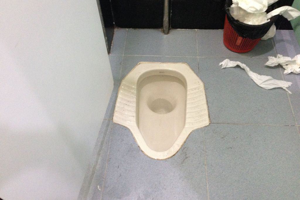 A traditional Chinese squat toilet low to the ground and with toilet paper falling out of a waste basket.