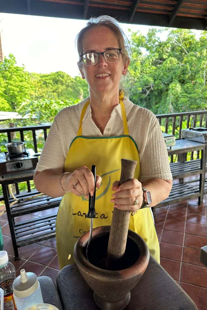 Me wearing a yellow apron and crushing the ingredients in the wooden mortar and pestle at the stainless steel worktop.