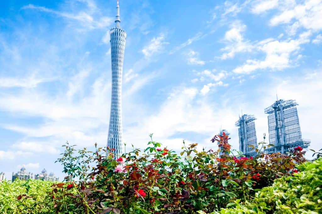 The Canton Tower in Guangzhou showing white against a bright blue sky with the city in the background and green trees in the foreground.