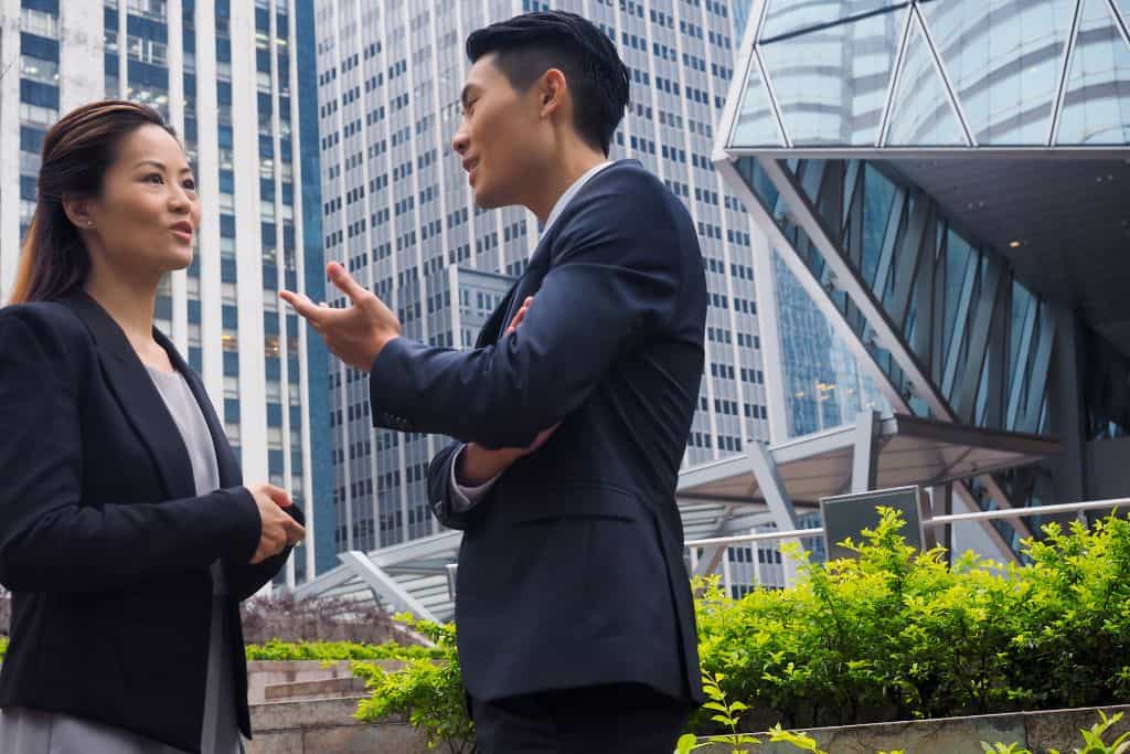 A man and a woman dressed in. business clothes having a discussion outside next to a modern building.