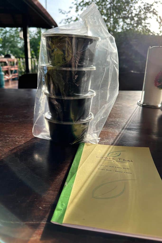 A stack of black plastic containers in a clear bag sitting on the table next to the yellow cookbook, ready for me to bring my leftovers home.