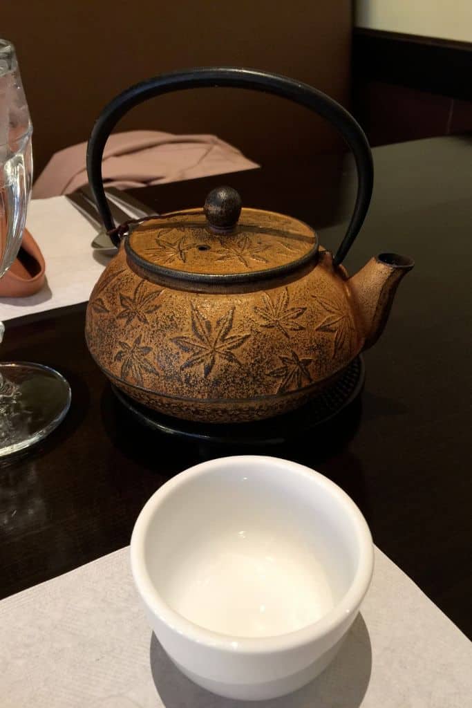 A small white tea cup in front of a heavy bronze colored Chinese tea pot.