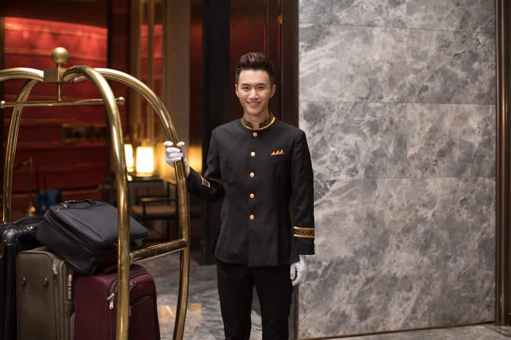 A young Chinese bellhop standing next to a luggage cart in a hotel.