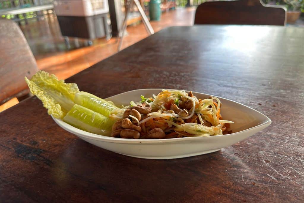 A freshly made papaya salad in a white dish sitting on the wooden table with extra nuts on top.